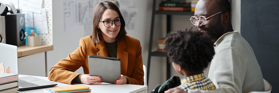 Female teacher sitting at a desk with young boy and his father during what appears to be a parent-teacher conference