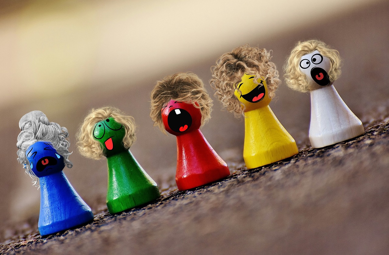 Six wooden game pieces are lined up side-by-side.  Each piece has a round, tapered base with a sphere at the top.  Each piece is a different color and has a different emoji expression on the sphere to create a "face" for the game mover.  All the movers have wigs, making them look more like people.