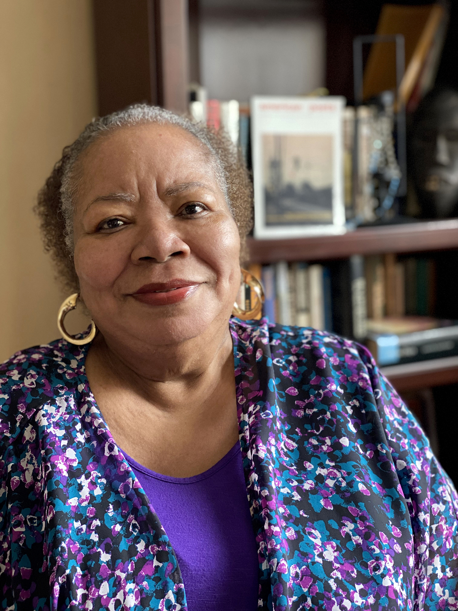 A photo of Angela Jackson.  An older, African-American woman, smiling and dressed in purple, is seated in front of a bookcase.  