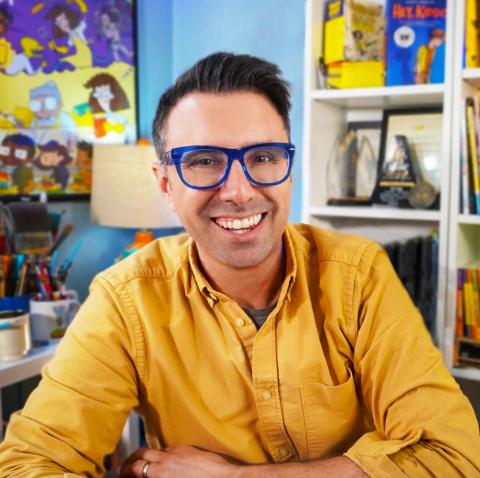 A man with dark short hair and glasses, seated at a desk or table, wearing a yellow button-down shirt.