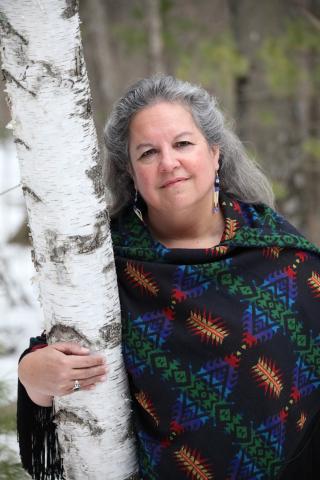 A woman with grey hair, wearing a poncho wrap, standing next to a birch tree.