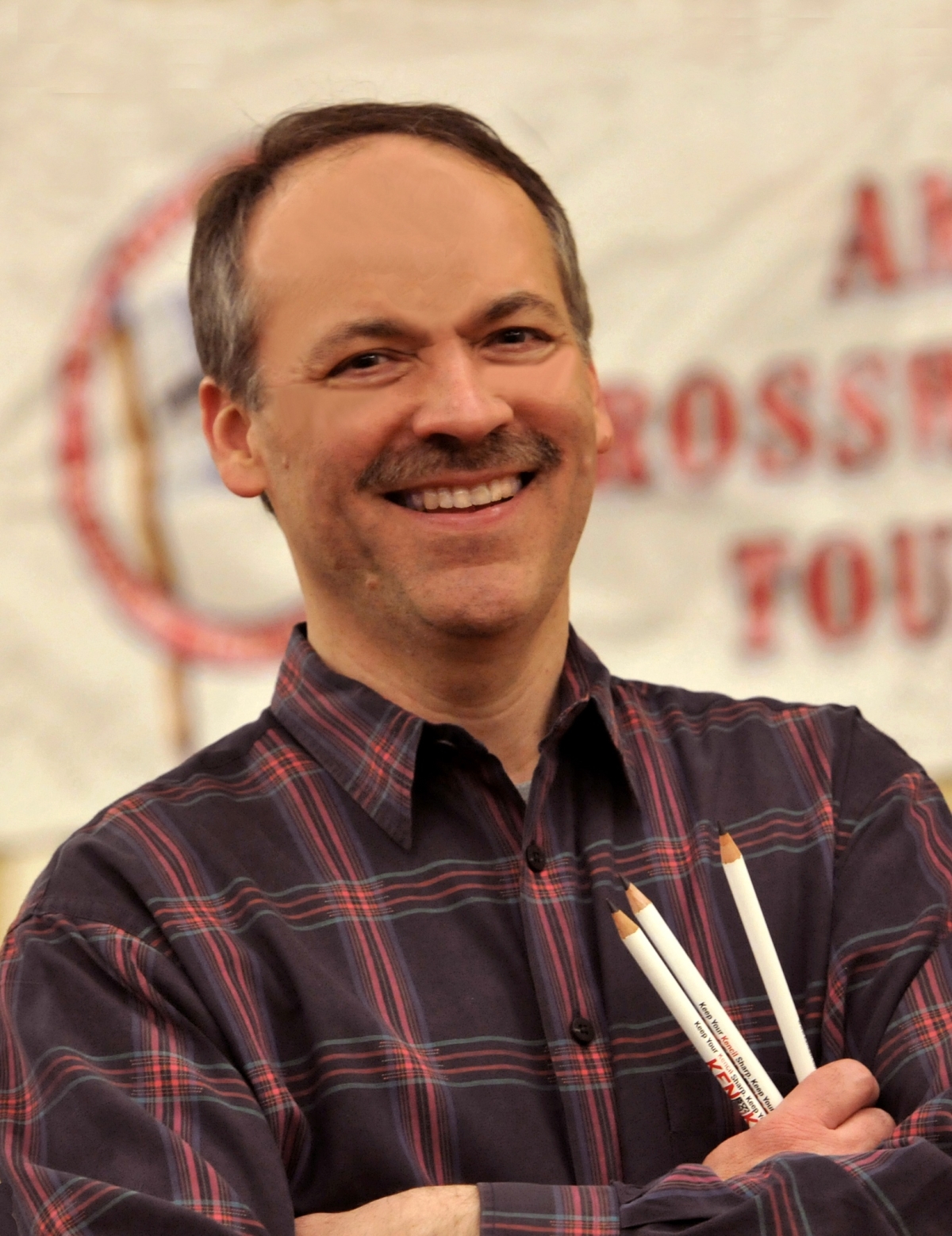 A smiling man with short hair and a mustache, wearing a plaid shirt and holding several pencils.