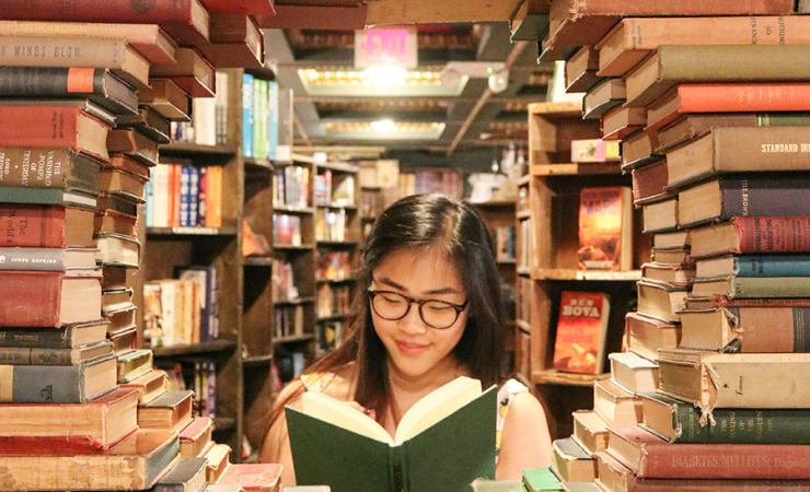 Teen girl in what appears to be a library surrounded by books with an open book in her hand